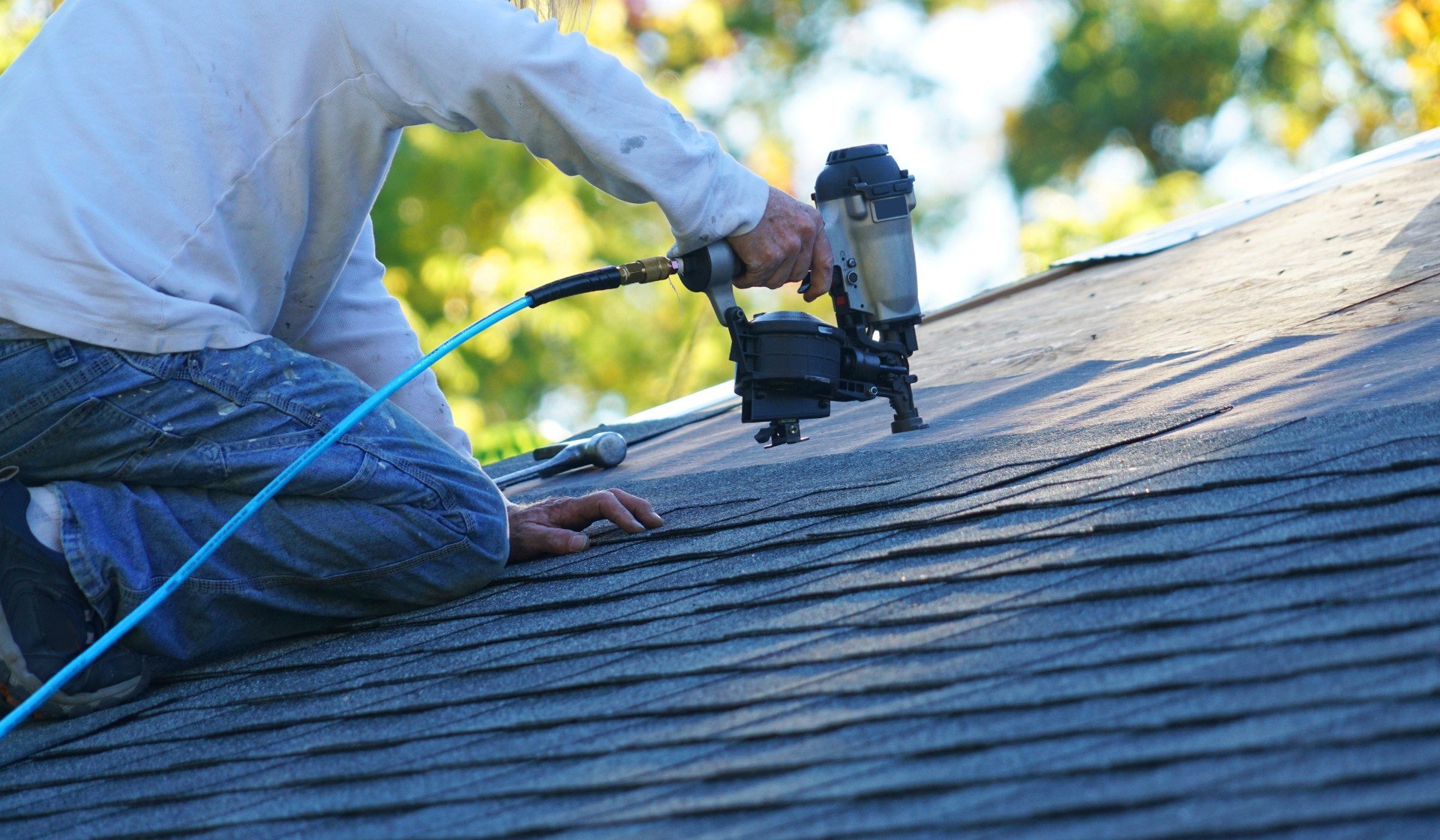 man kneels on roof while using nail gun to attach shingles